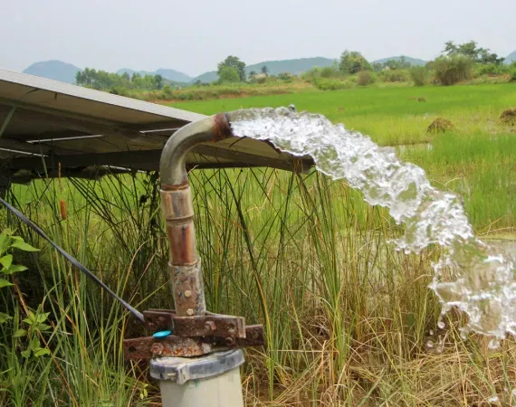 Water pours from a pipe powered by solar panels in a rural field with hills in the background.