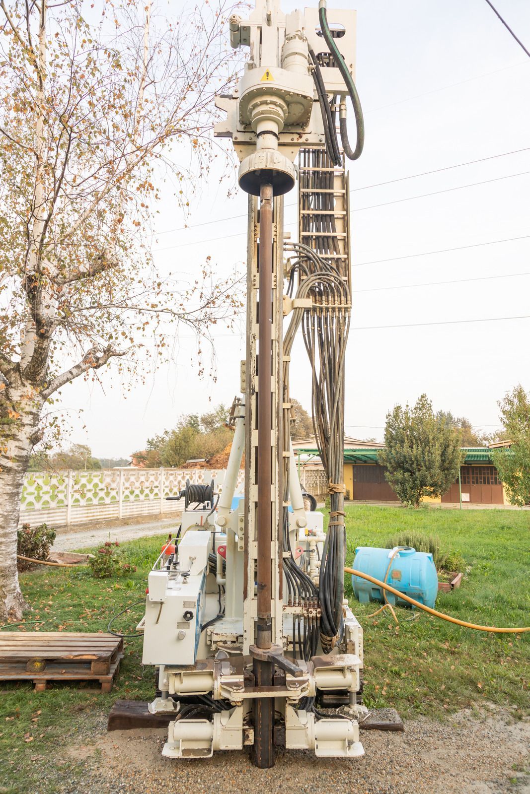 A white vertical drilling rig stands outdoors on grass near a tree and a blue water tank.