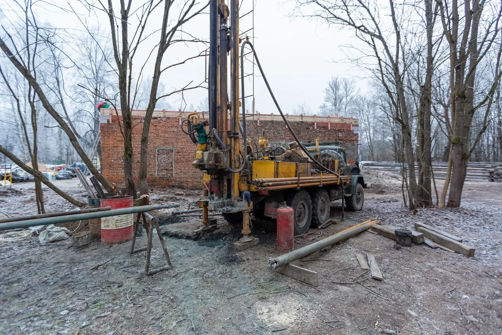 A yellow truck-mounted drilling rig operates on a snowy, wooded site in front of a weathered brick building.