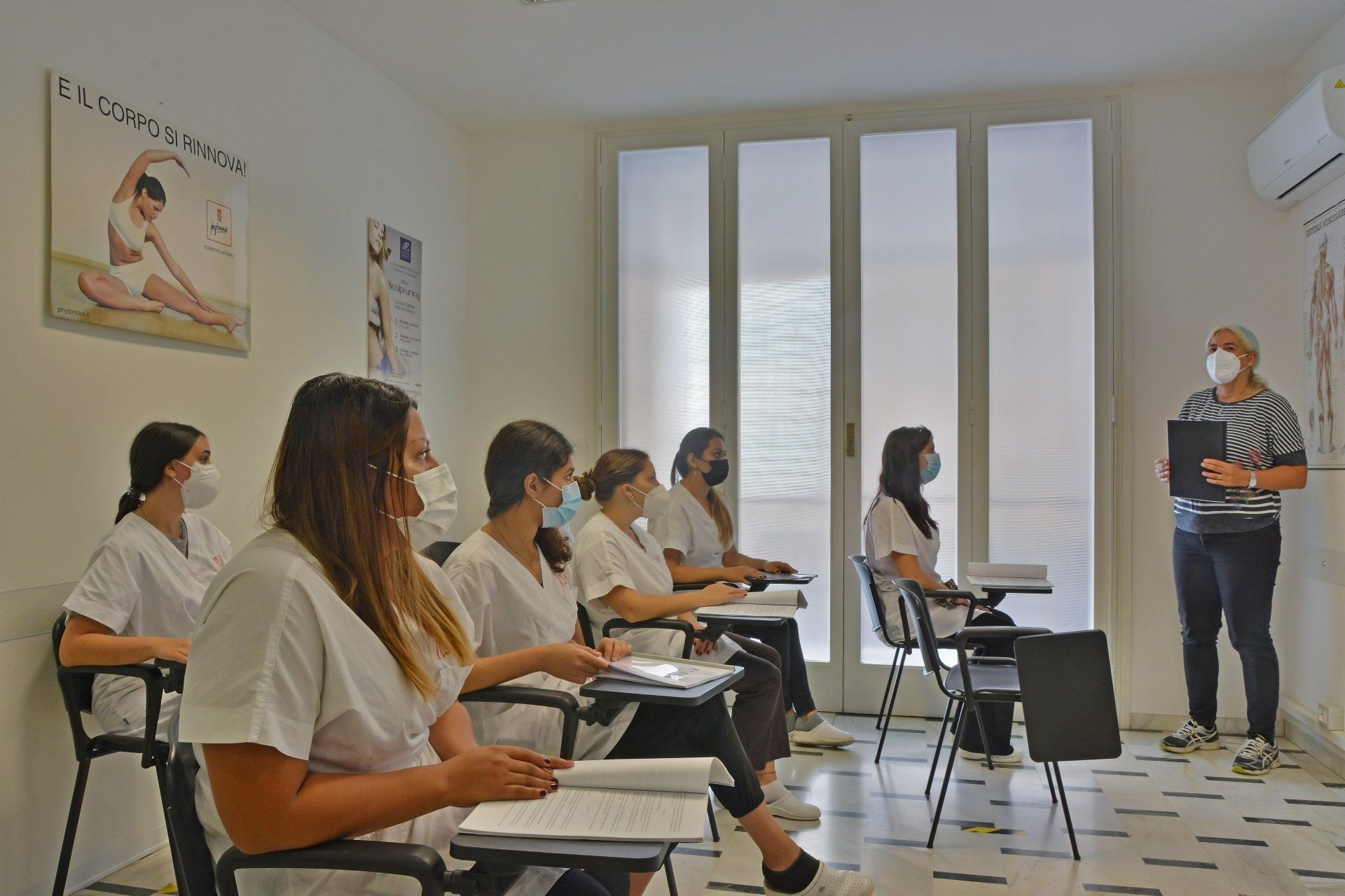 Impostazione della classe: un insegnante con la mascherina tiene lezione agli studenti in camice bianco, alcuni dei quali indossano la mascherina.