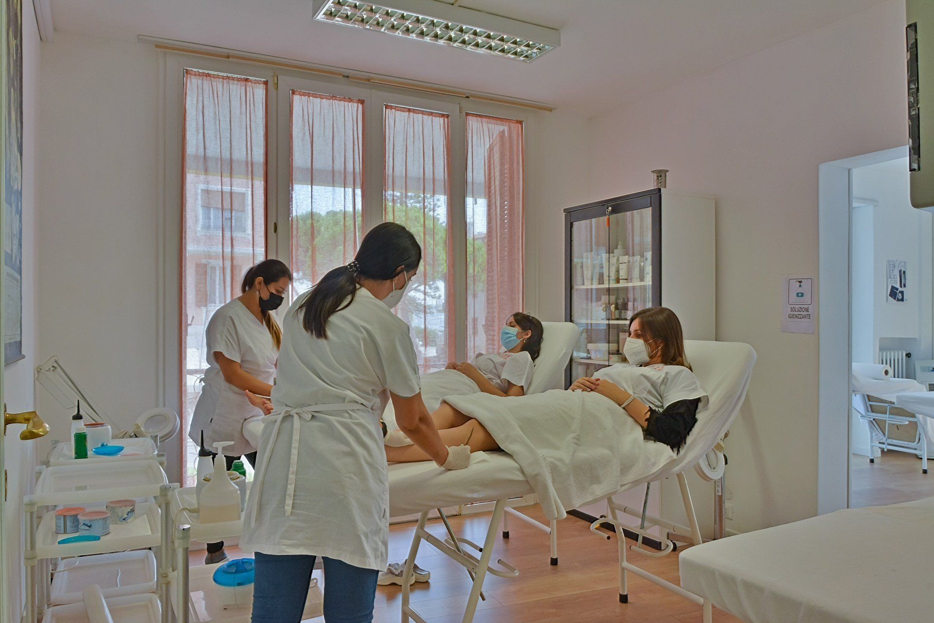 Persone in una clinica: due pazienti sui letti, due membri dello staff che li assistono, eseguono procedure mediche. Stanza luminosa.