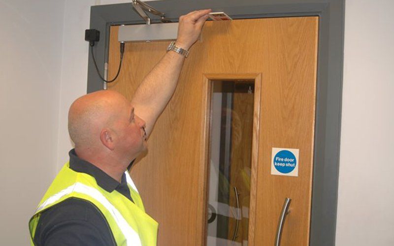 Man in a yellow vest inspecting a door with an overhead closer. Fire exit sign visible.