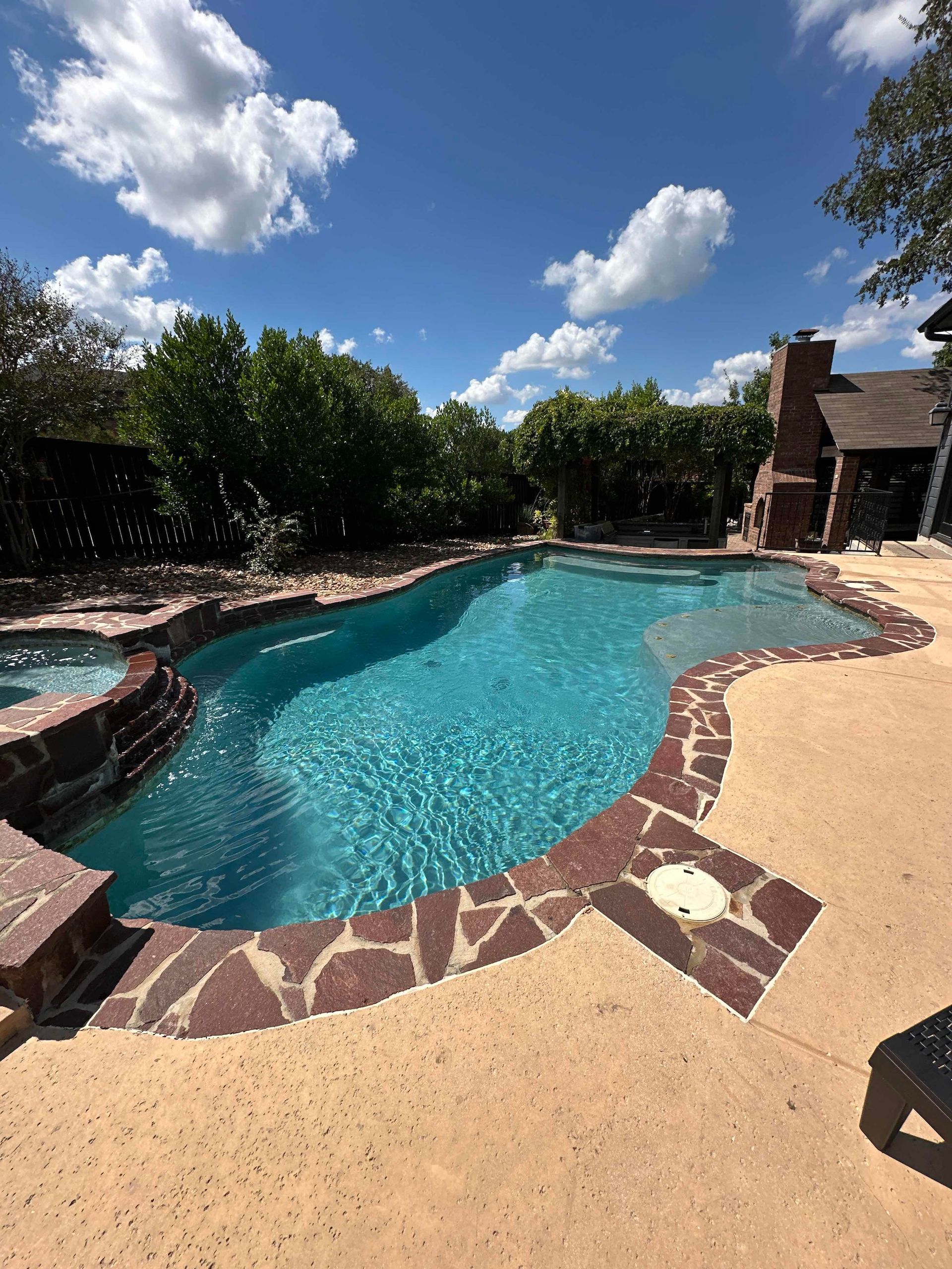 Backyard swimming pool with blue water, stone deck, trees, and a bright cloudy sky
