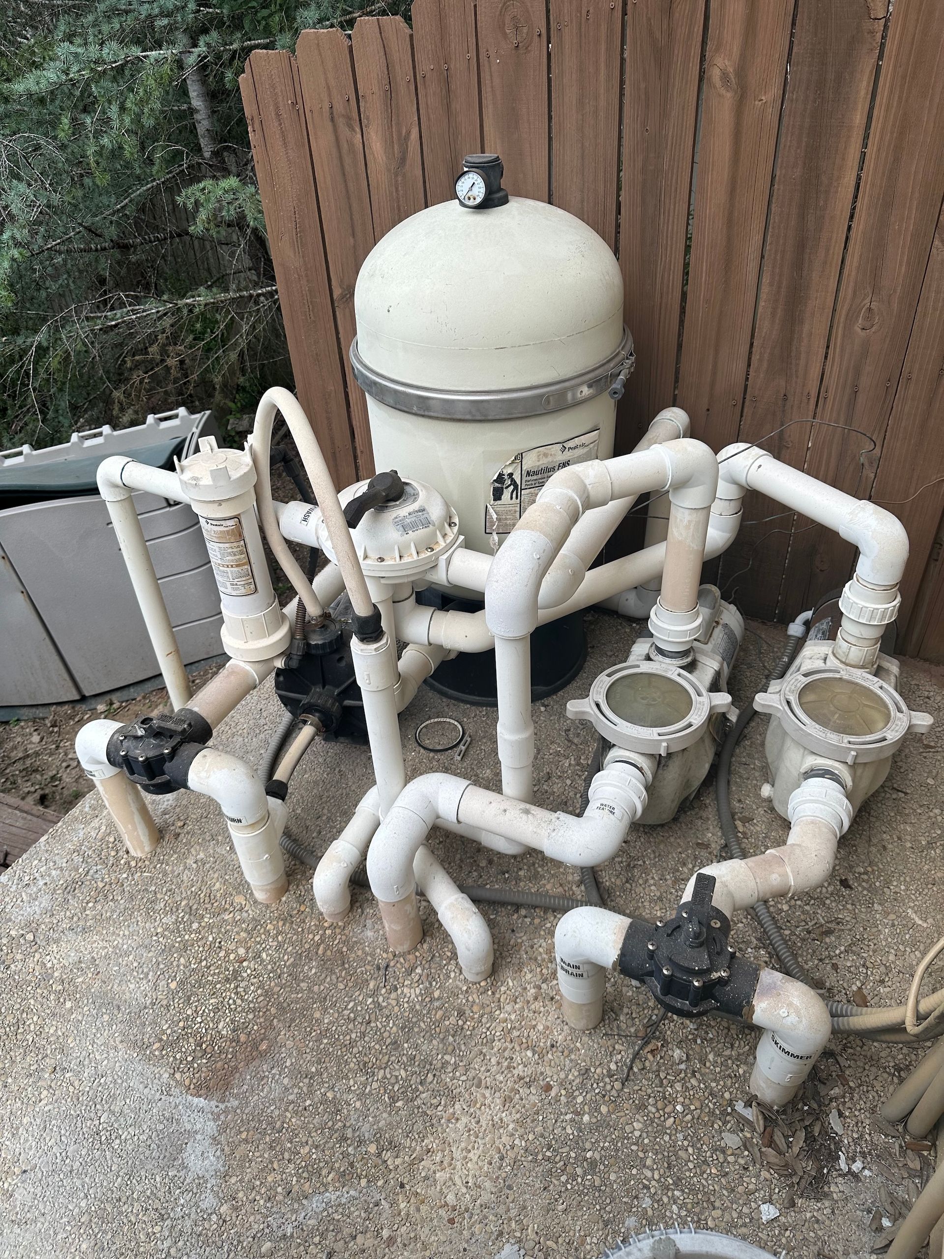 Pool filter and plumbing system with white pipes and a beige tank beside a wooden fence.