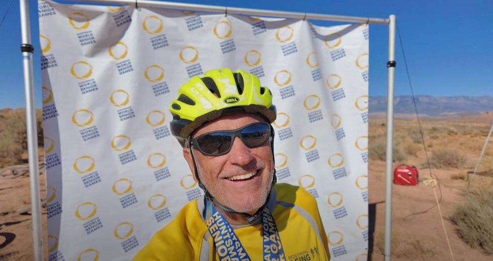 Smiling cyclist in yellow gear and helmet posing outdoors before a race banner in a desert setting