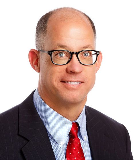 Portrait of a man in glasses, dark suit, blue shirt, and red tie against a white background