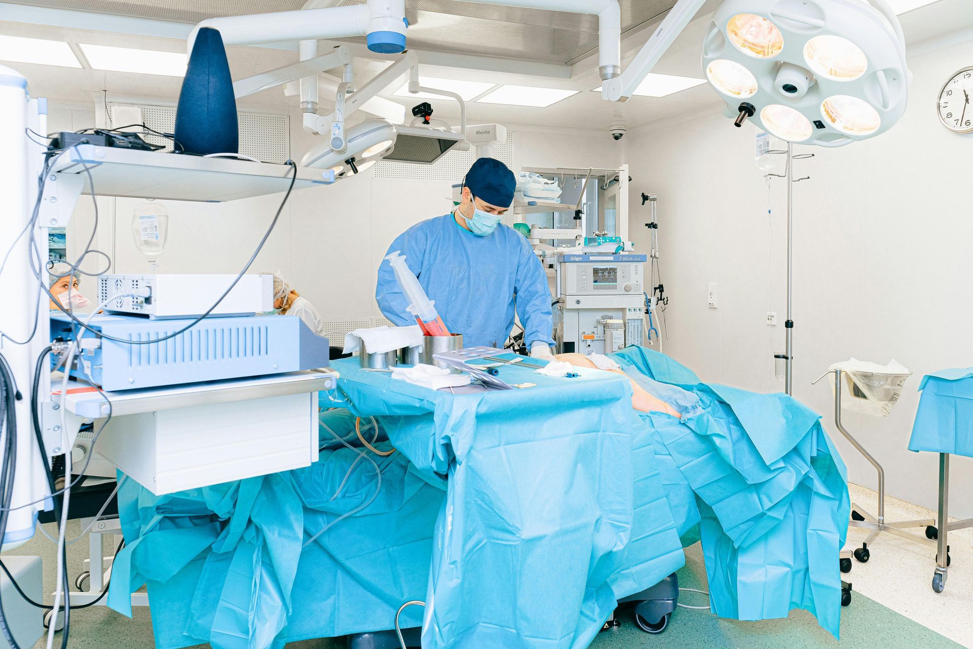 Surgeon in blue scrubs operating in a bright hospital operating room with surgical equipment and draped table.