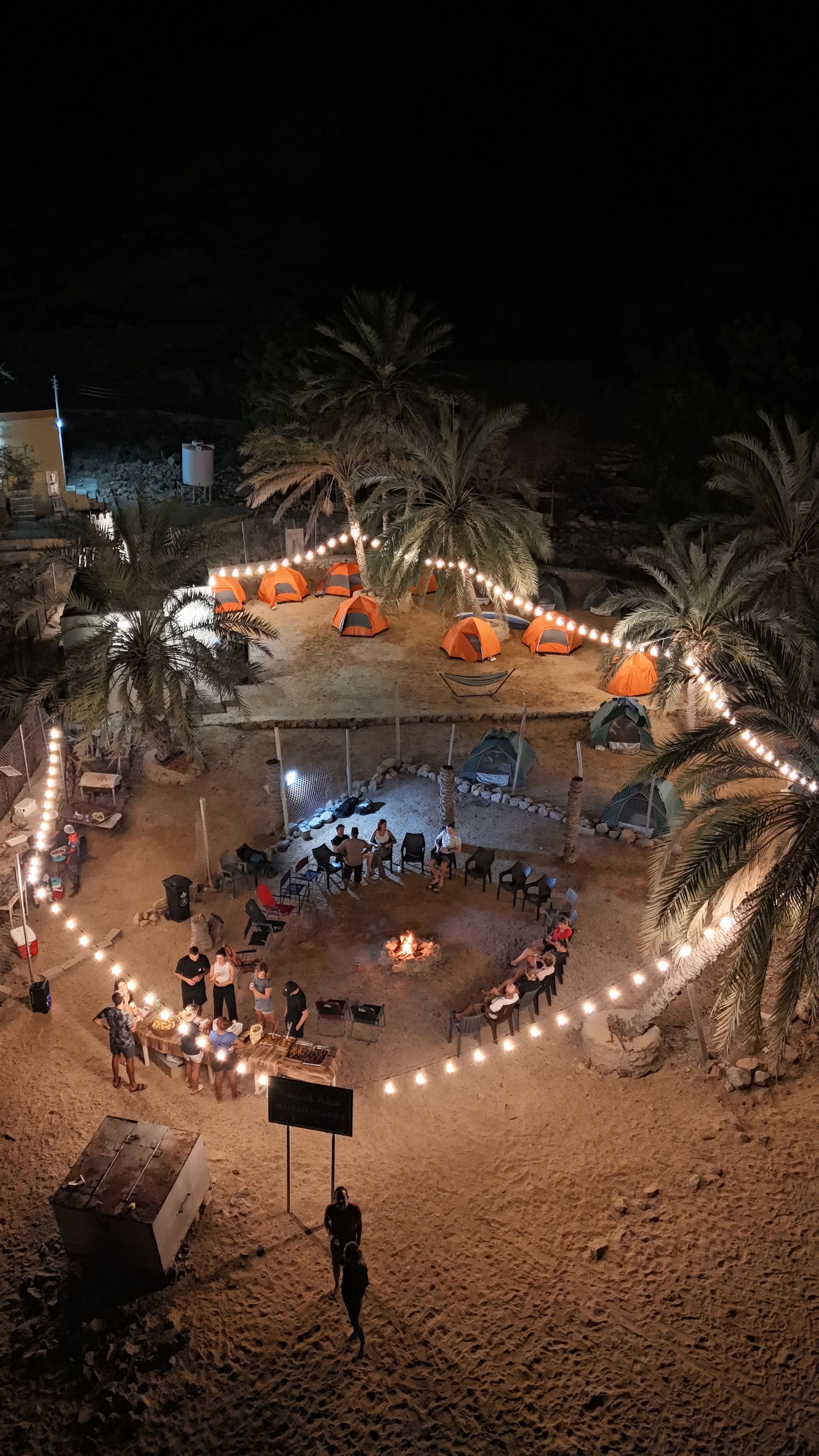 Aerial view of a nighttime beach gathering with a lit circular seating area, palm trees, and tents in Dibba Musandam.