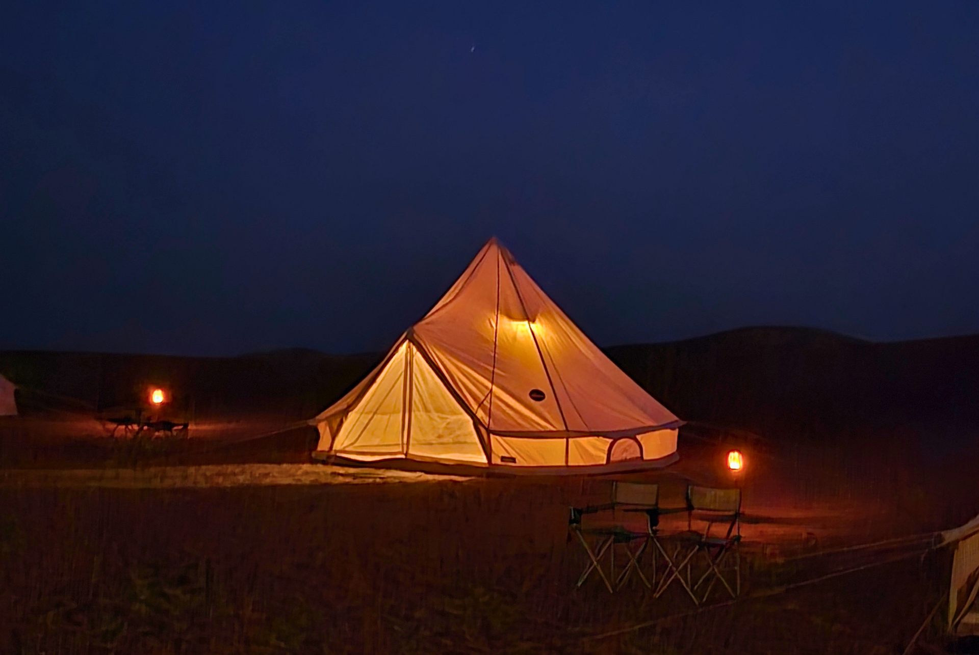 Lit glamping tent glowing at night in a dark desert campsite with a chair and lanterns in Al Quaa.