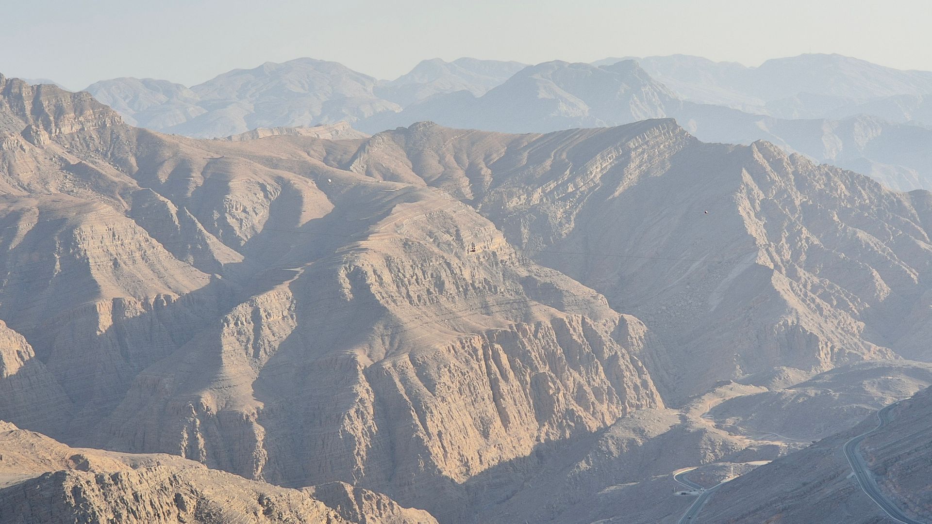 Rocky mountain ridges under a hazy sky, with layered tan and gray peaks in the distance in Jebel Jais