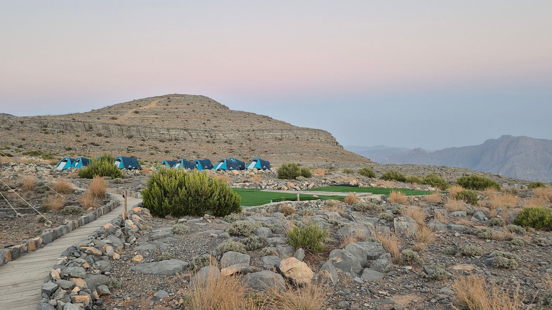 Rocky hillside camp with blue tents at dusk under a pink sky in Jebel Jais' peak.