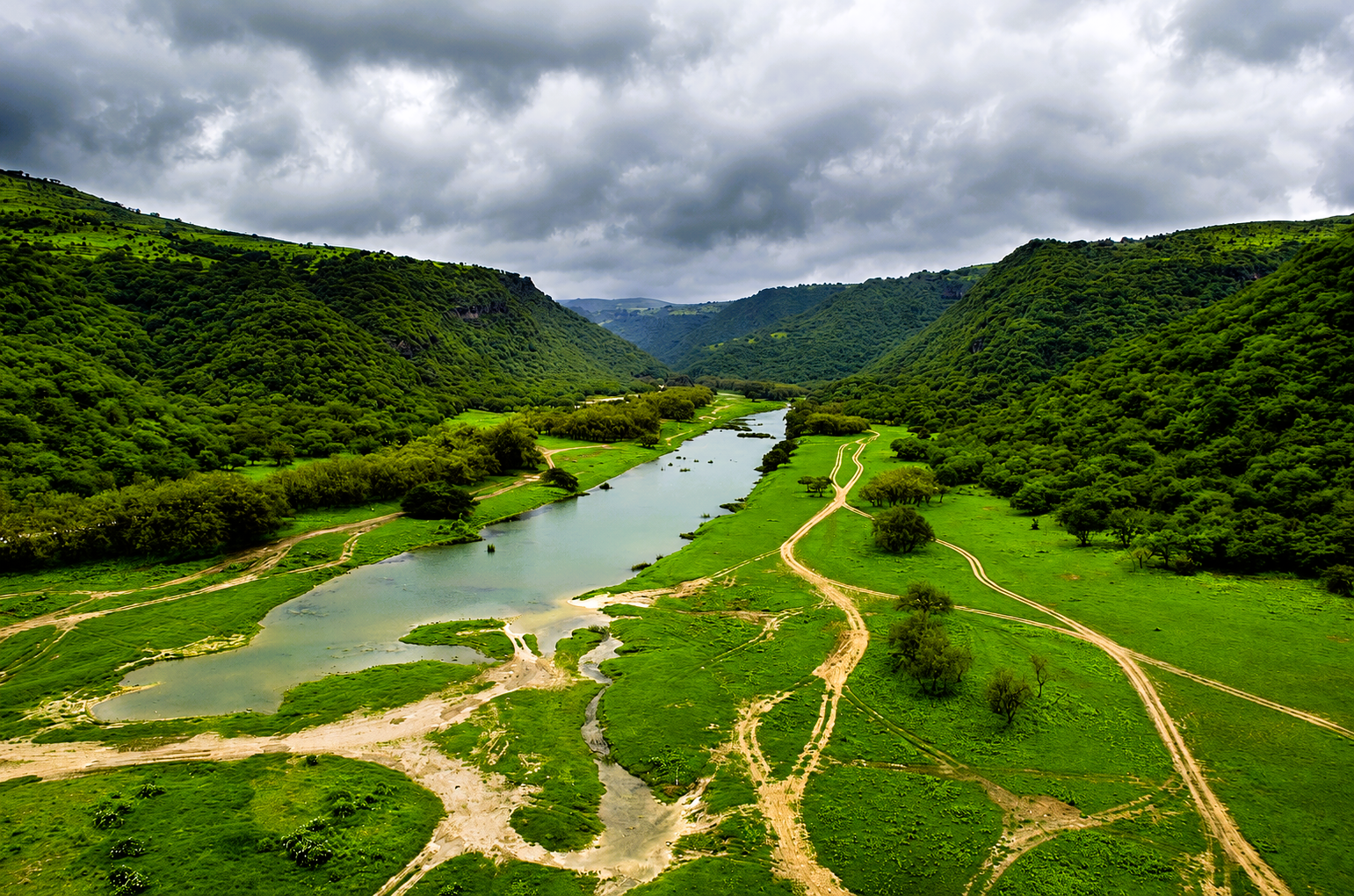 erial view of Wadi Darbat in Salalah during the Khareef season, featuring a winding river, lush green valley, and dramatic cloud-covered mountains
