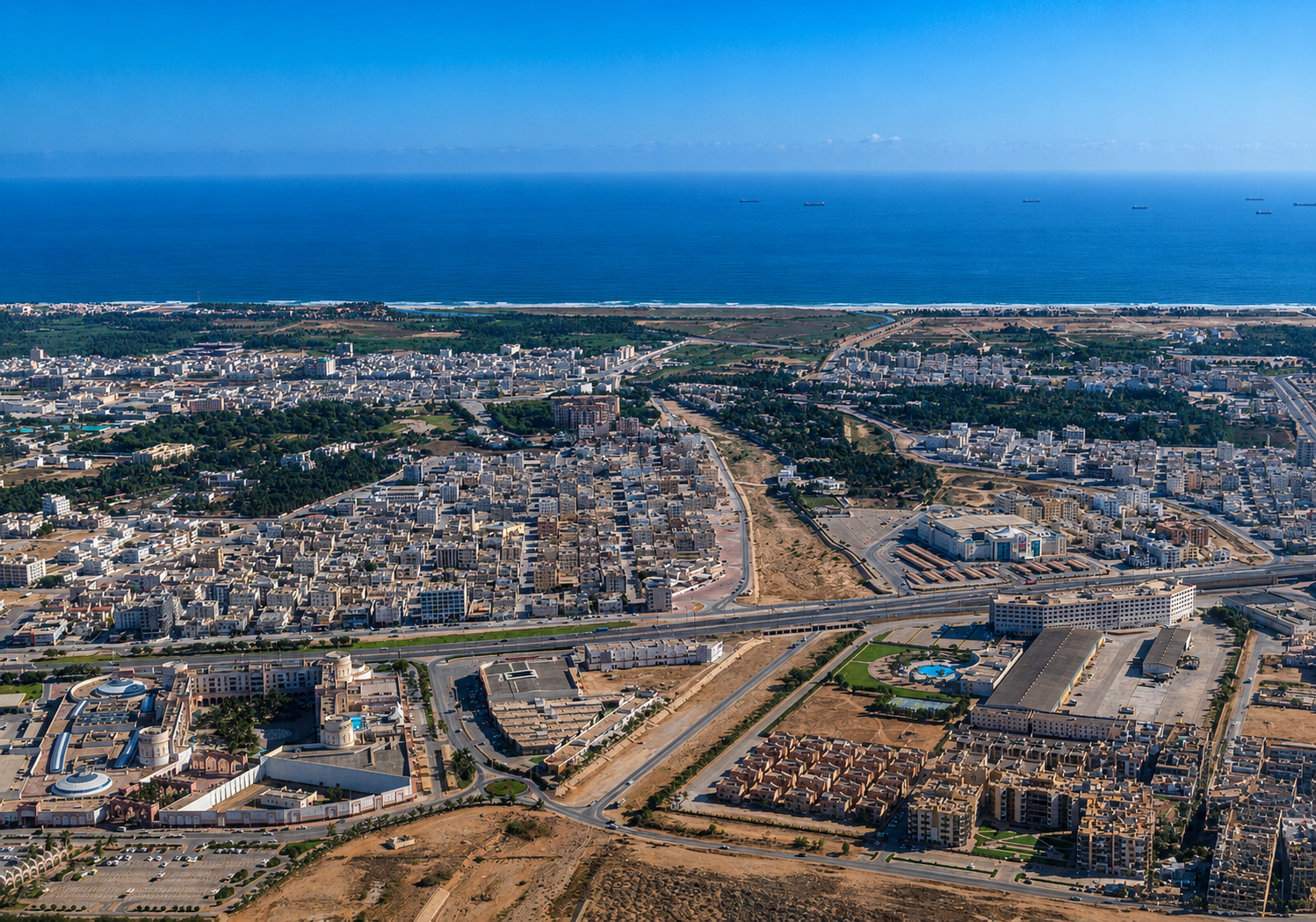 Aerial view of a coastal city with roads, buildings, and the ocean under a blue sky in Salalah.