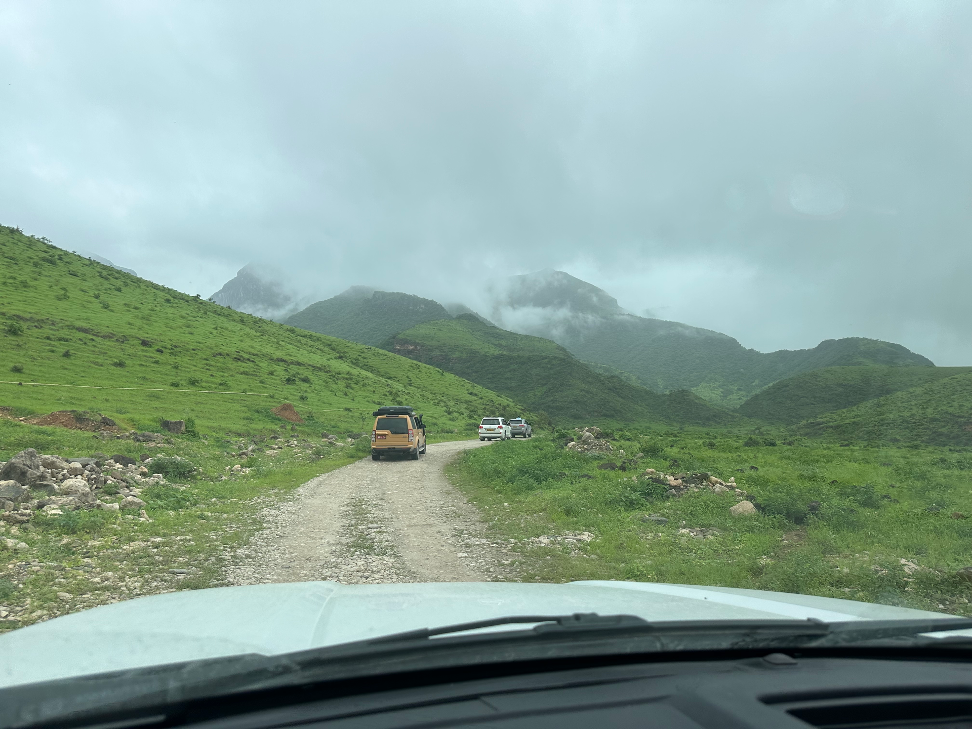 SUVs driving on a gravel road through green mountains under cloudy skies towards a secret waterfall in Salalah.
