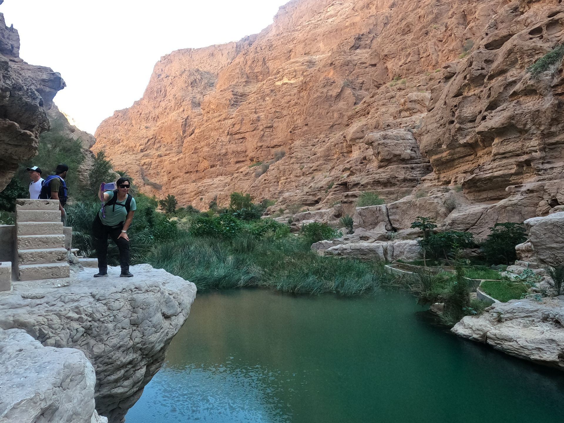 Two hikers standing by a turquoise river in a rocky canyon in Wadi Shab in Oman.