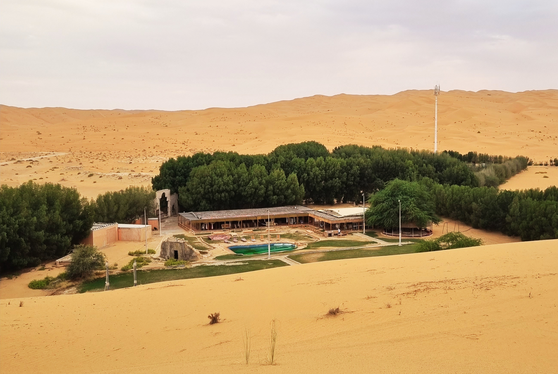 Desert oasis with a small farm, green trees, and a turquoise pool beneath sandy dunes in Liwa. 