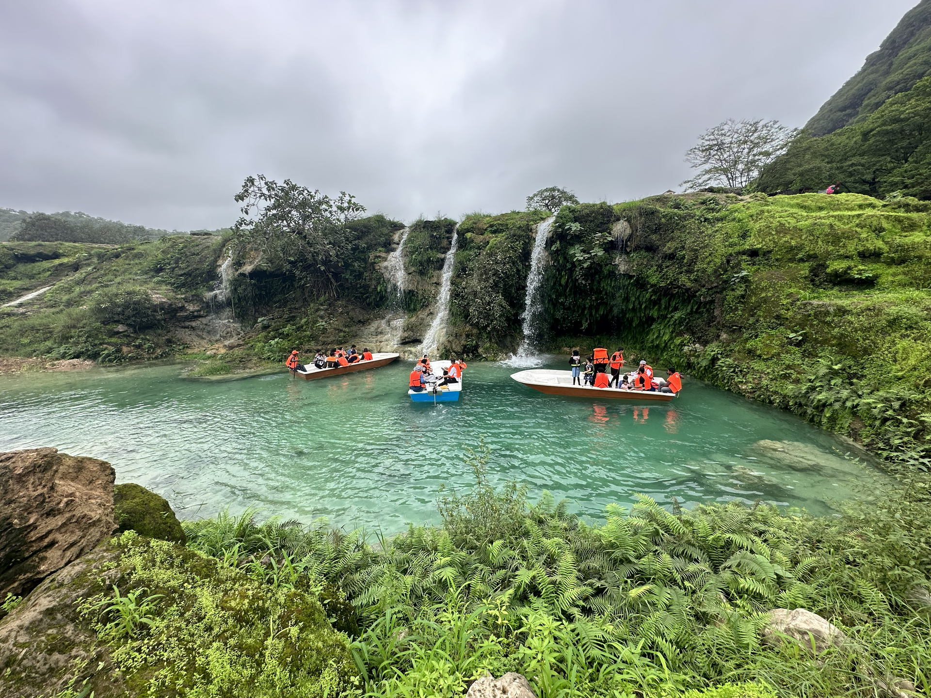 Kayakers in red life vests beneath a small waterfall in a turquoise rocky pool in Wadi Darbat, Salalah.
