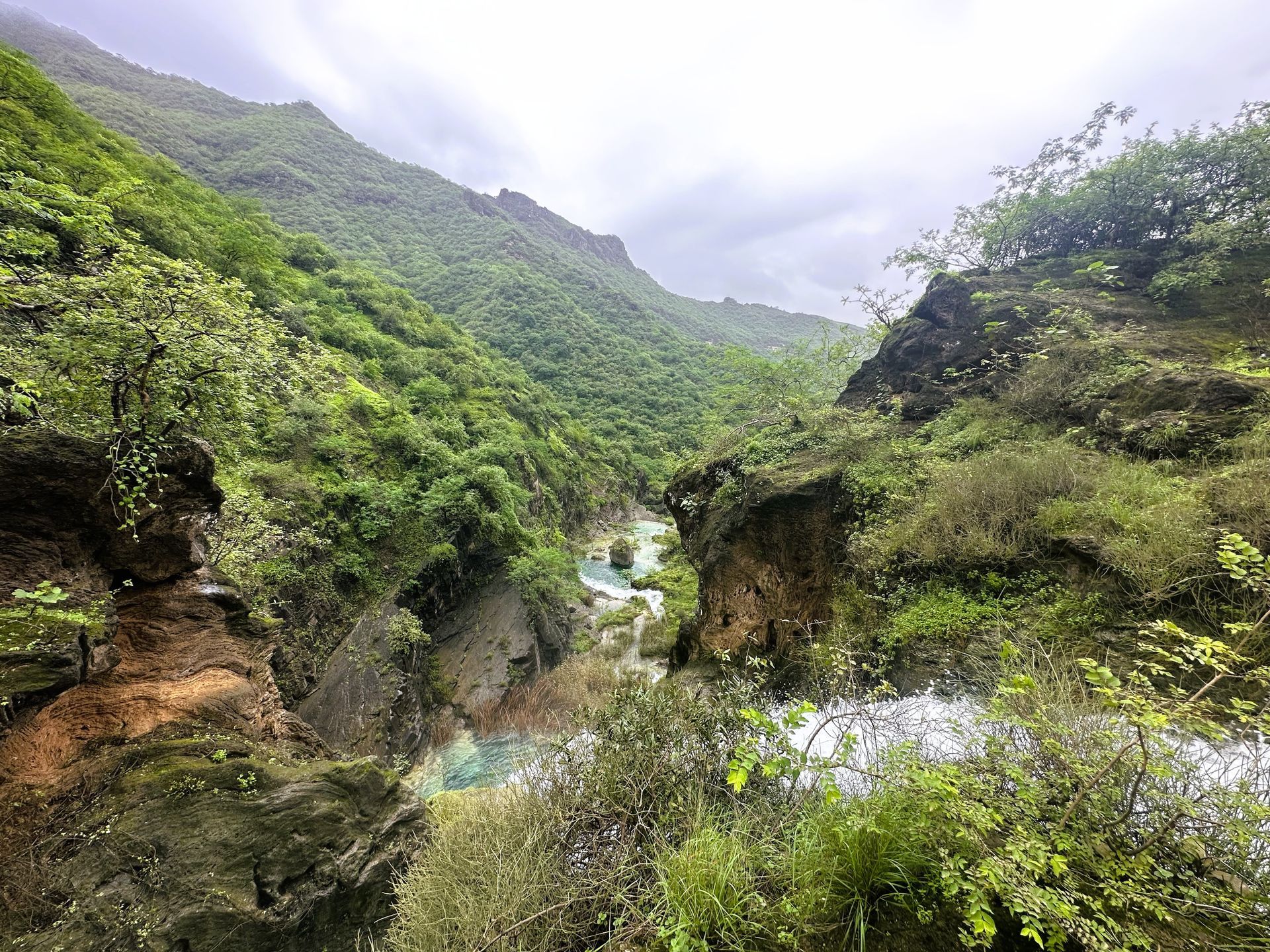 Lush green mountain valley with rocky cliffs and a winding stream under a cloudy sky in the hidden waterfall in Salalah.
