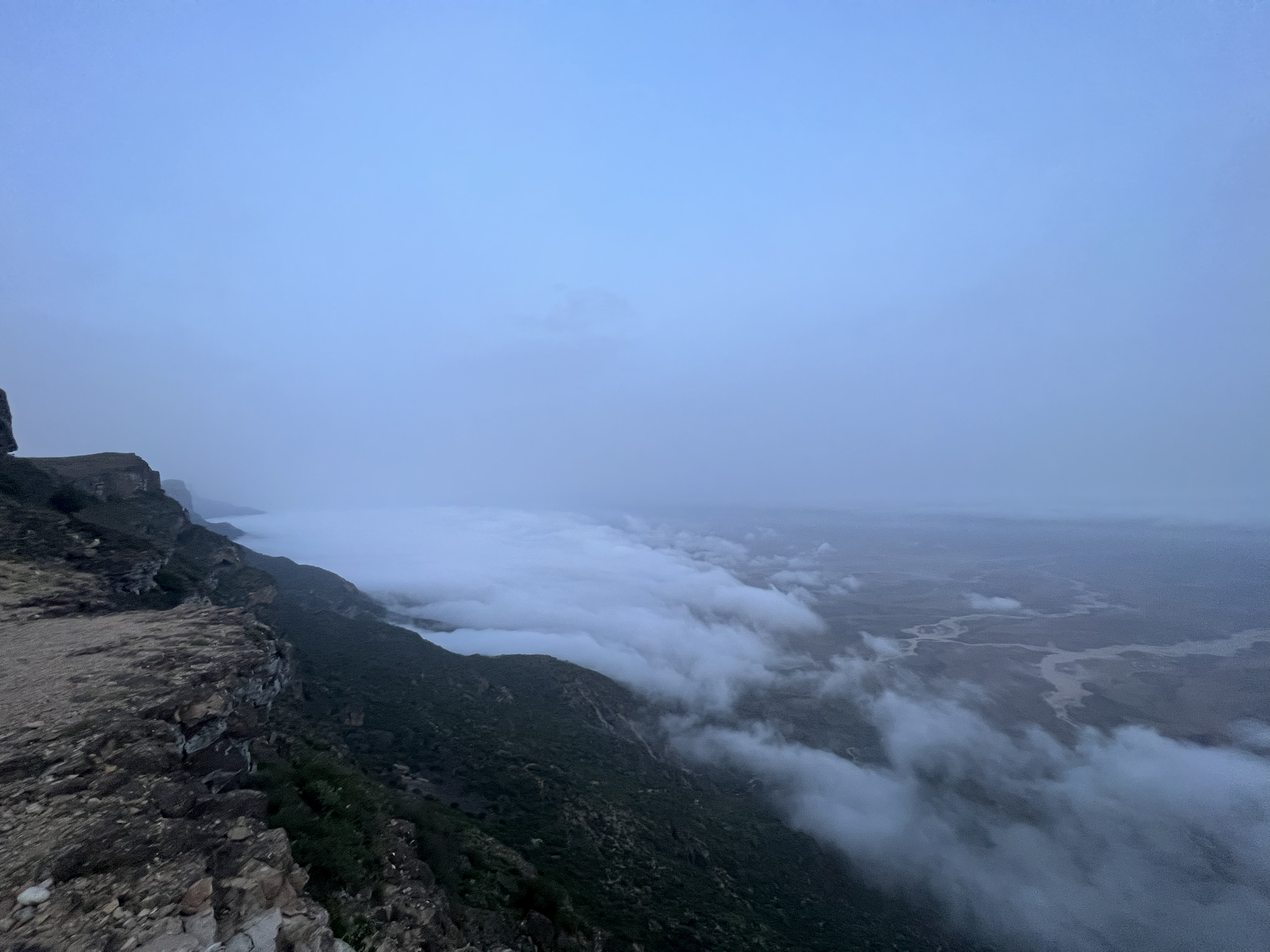 Foggy mountain overlook above a vast valley under a blue sky in Jebel Samhan.