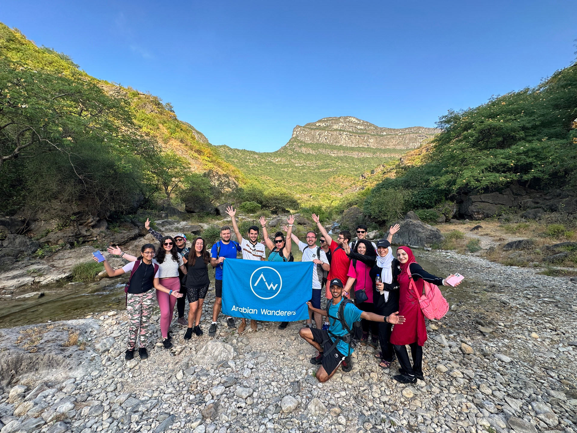 Group of people on a rocky trail holding Arabian Wanderers' flag in Salalah with green mountains and blue sky behind them
