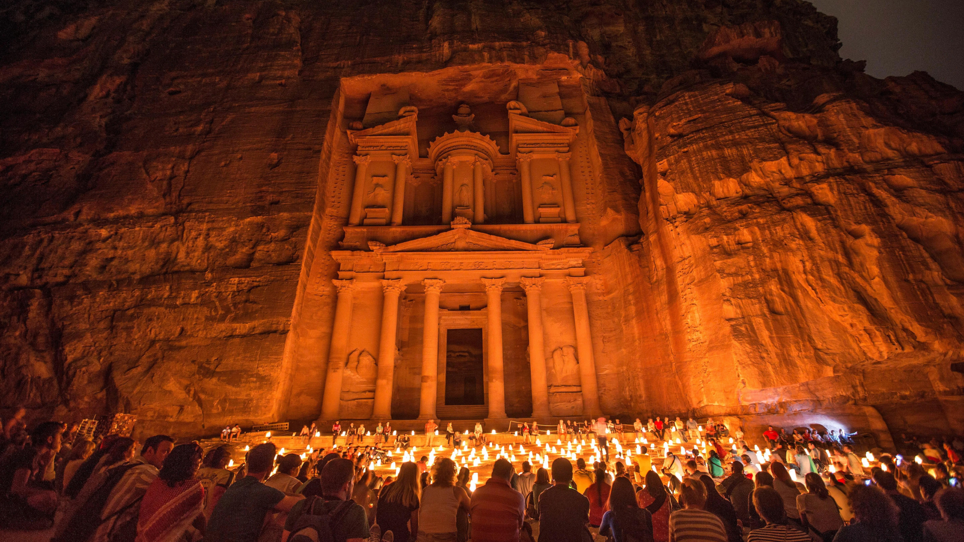 Crowd with candles before the illuminated Petra Treasury carved into a sandstone cliff at night.