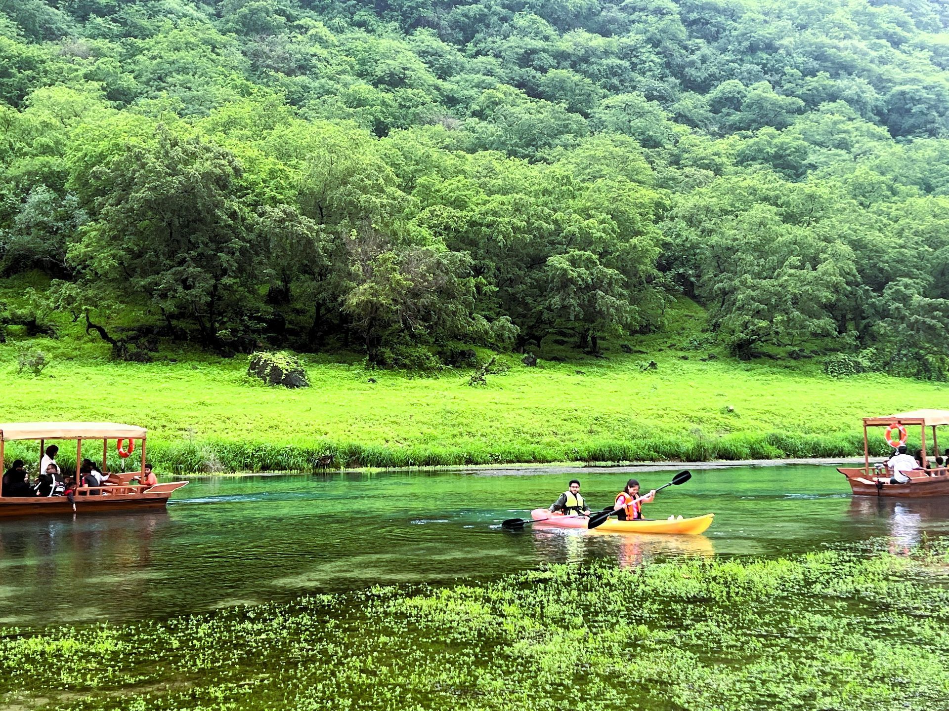 Boats with people on a calm river beside lush green forested hills in Wadi Darbat.