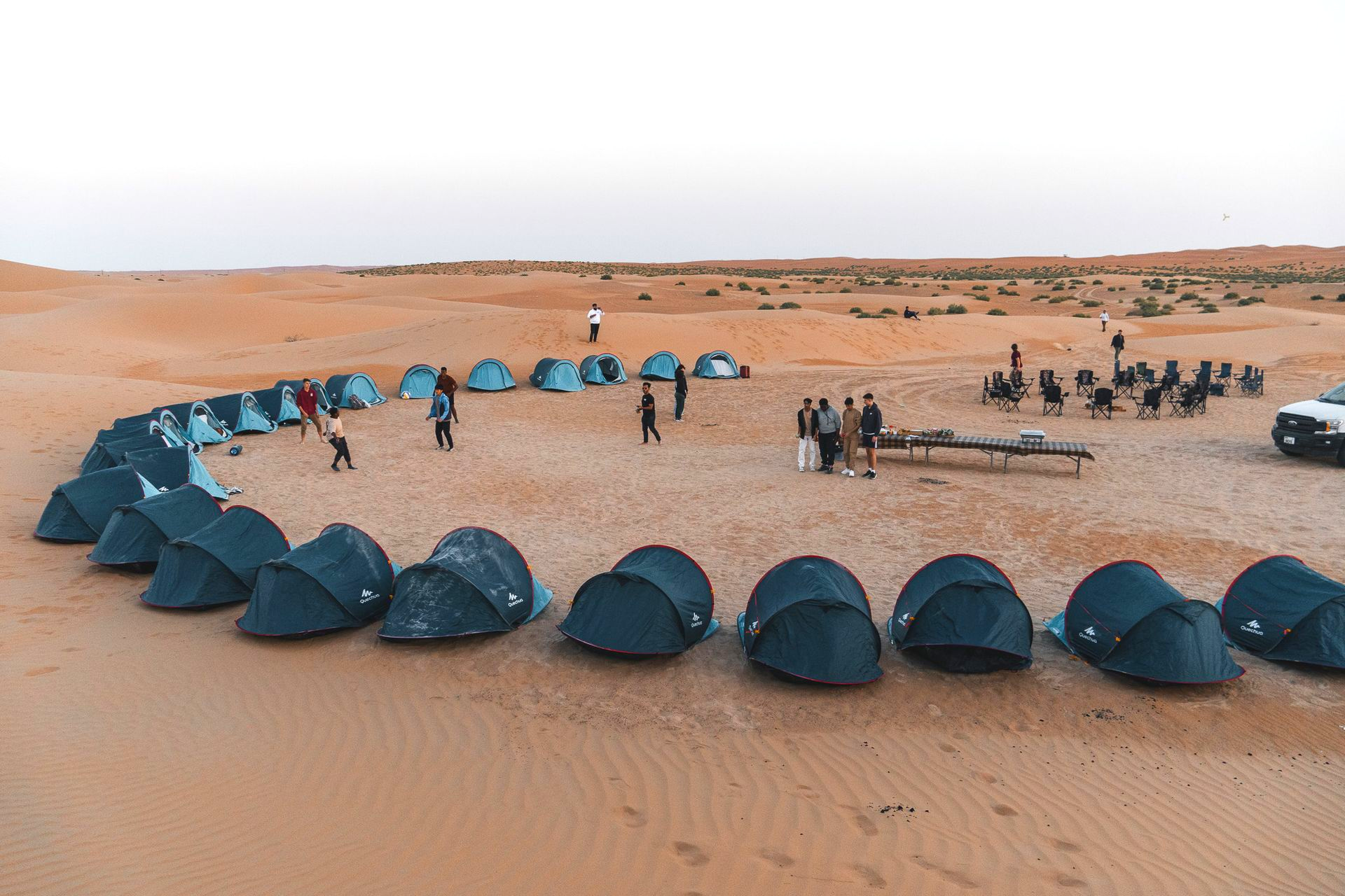 Blue tents and people arranged in a line across a sandy desert campsite in Al Quaa.