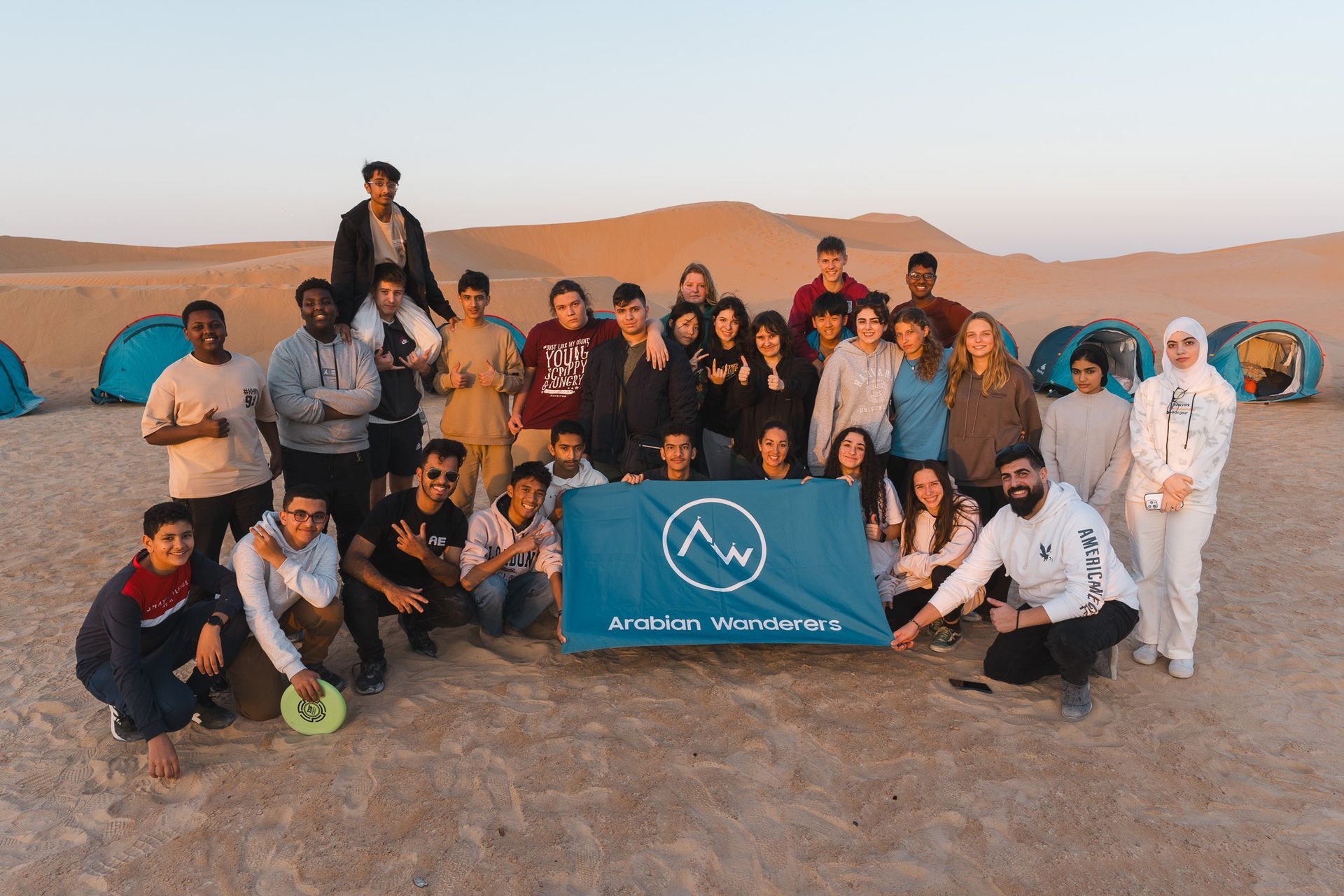 Students posing outdoors at sunset behind a blue Arabian Wanderers flag in a desert landscape in Al Quaa.