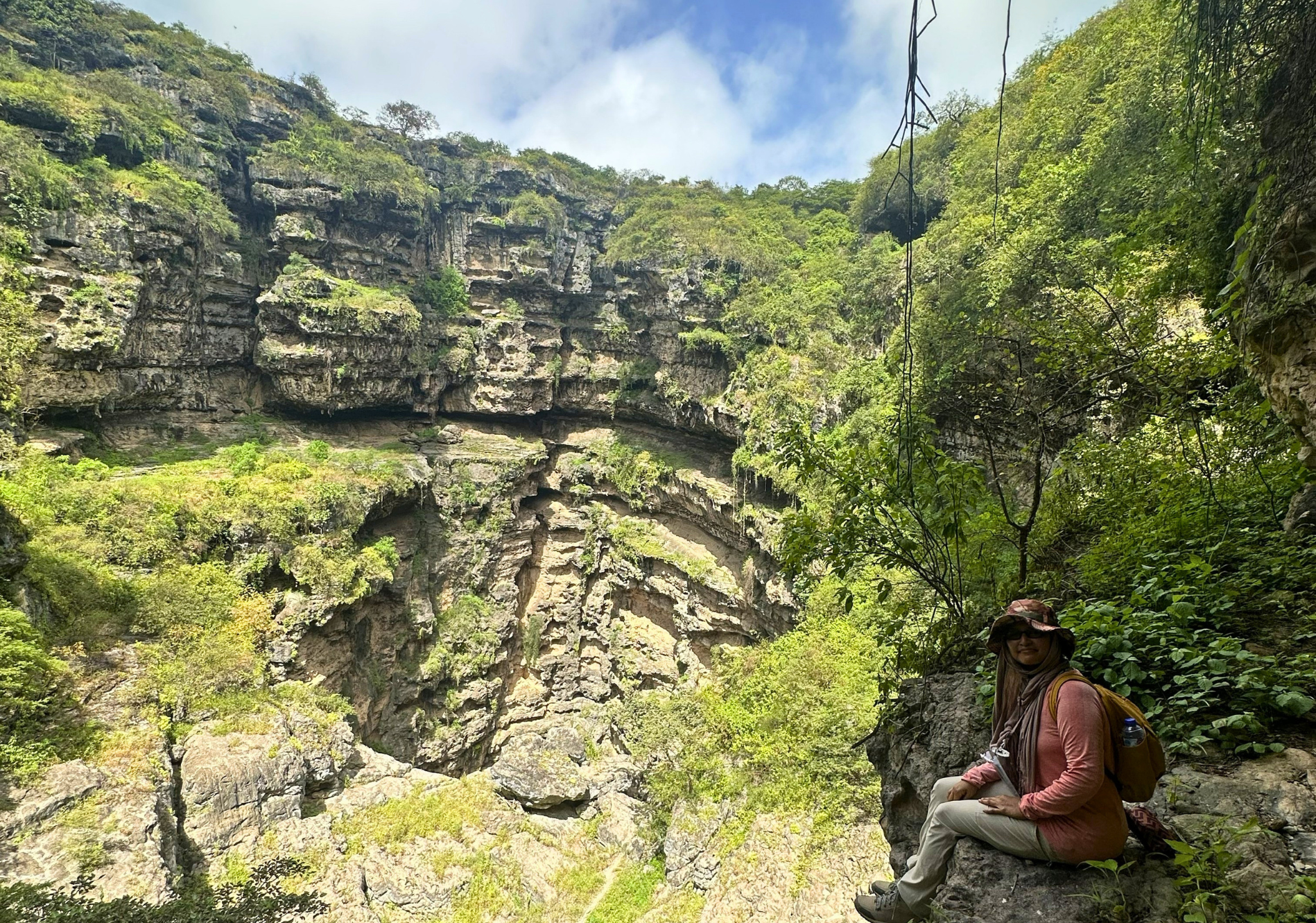 Person sitting on a rock ledge in a lush canyon beneath layered cliffs and blue sky in Tawi Atair Sinkhole.