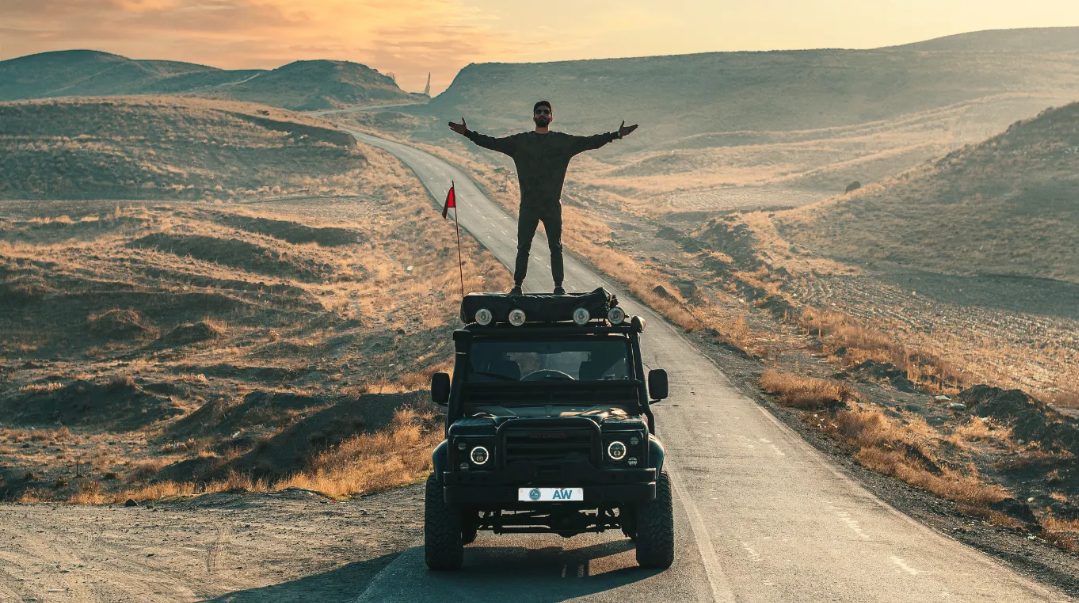 Person standing through a vehicle sunroof on a dirt road in a desert landscape, arms outstretched.
