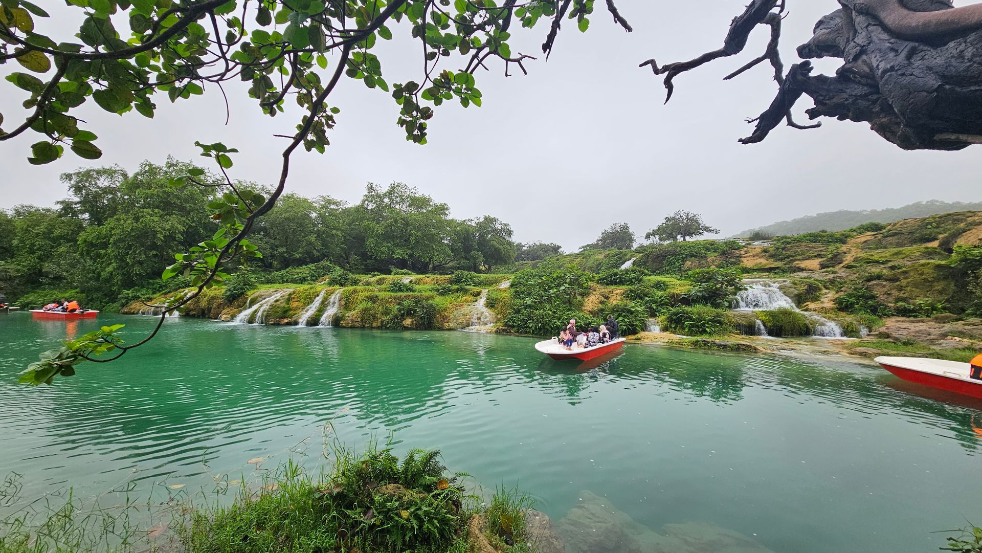 Serene green lake with small boats, waterfalls, and lush trees under a cloudy sky in Wadi Darbat, Salalah.