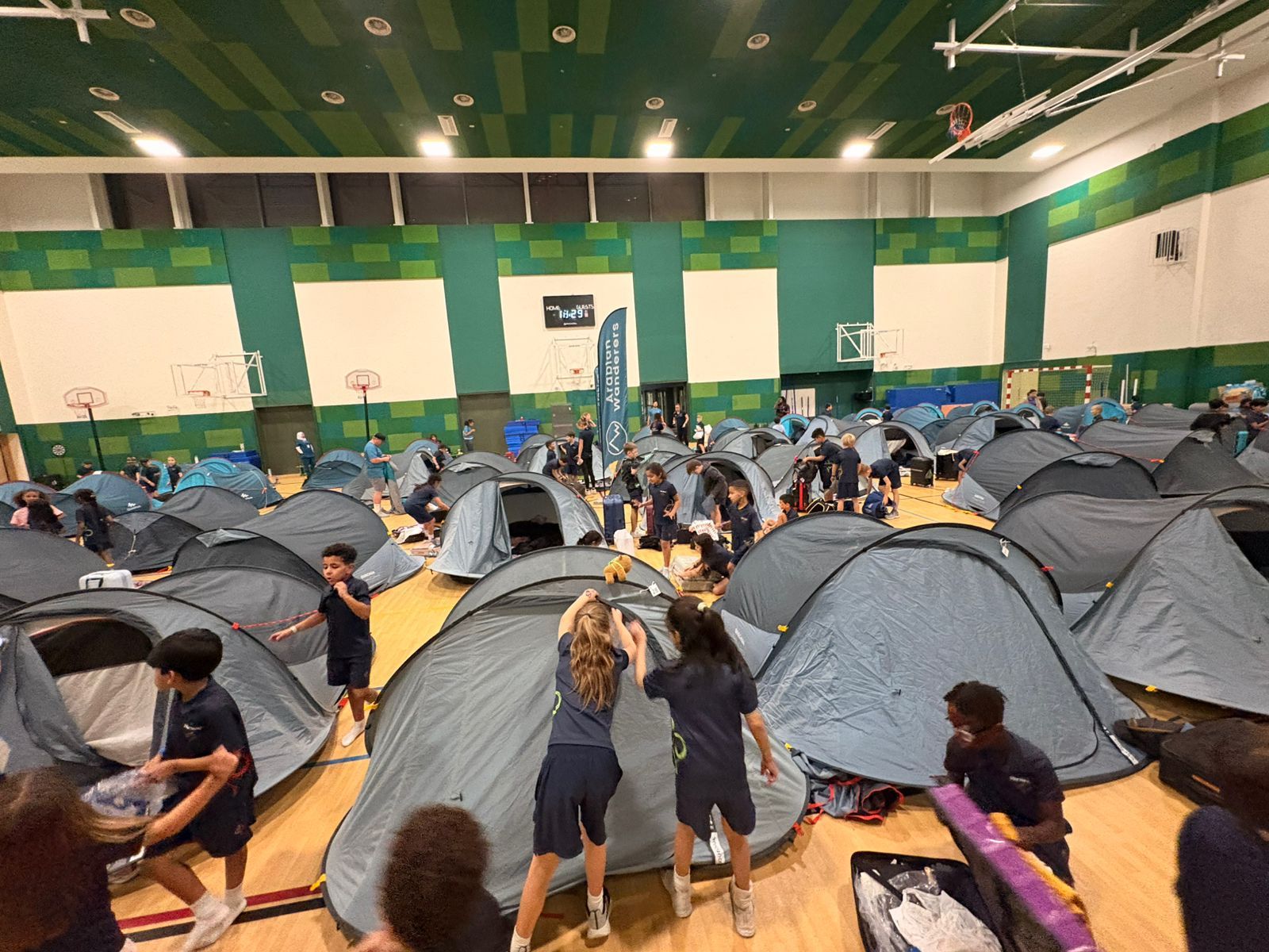 Students setting up tents in a gymnasium with green walls and a wooden floor in Hartland International School.
