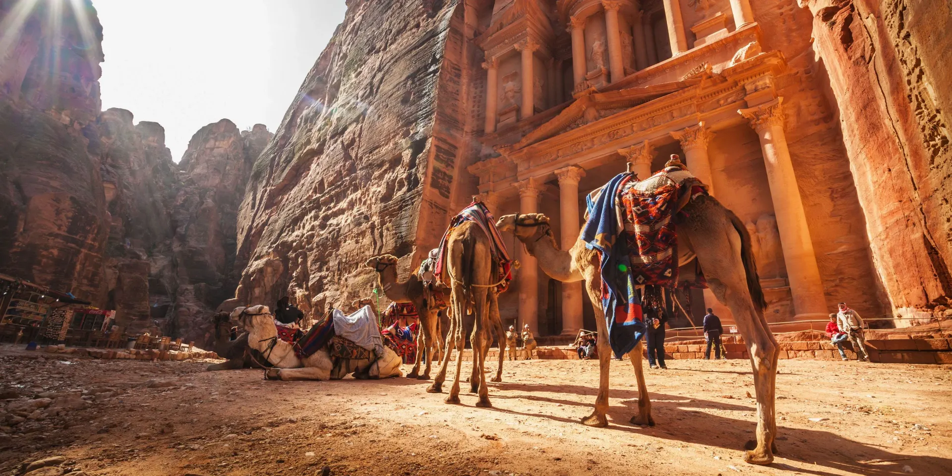 Camels and people walking through a red sandstone canyon with towering carved cliffs in Petra.