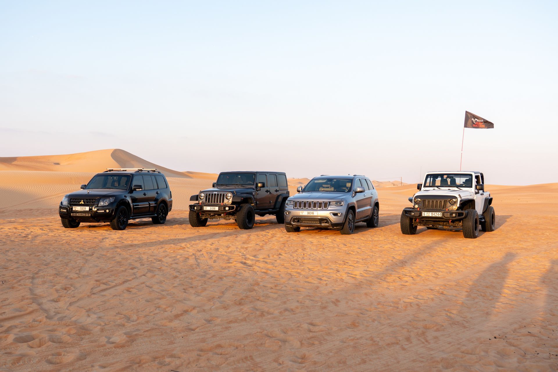 Four off-road vehicles parked on desert sand dunes, one flying a flag at sunset in Al Quaa.