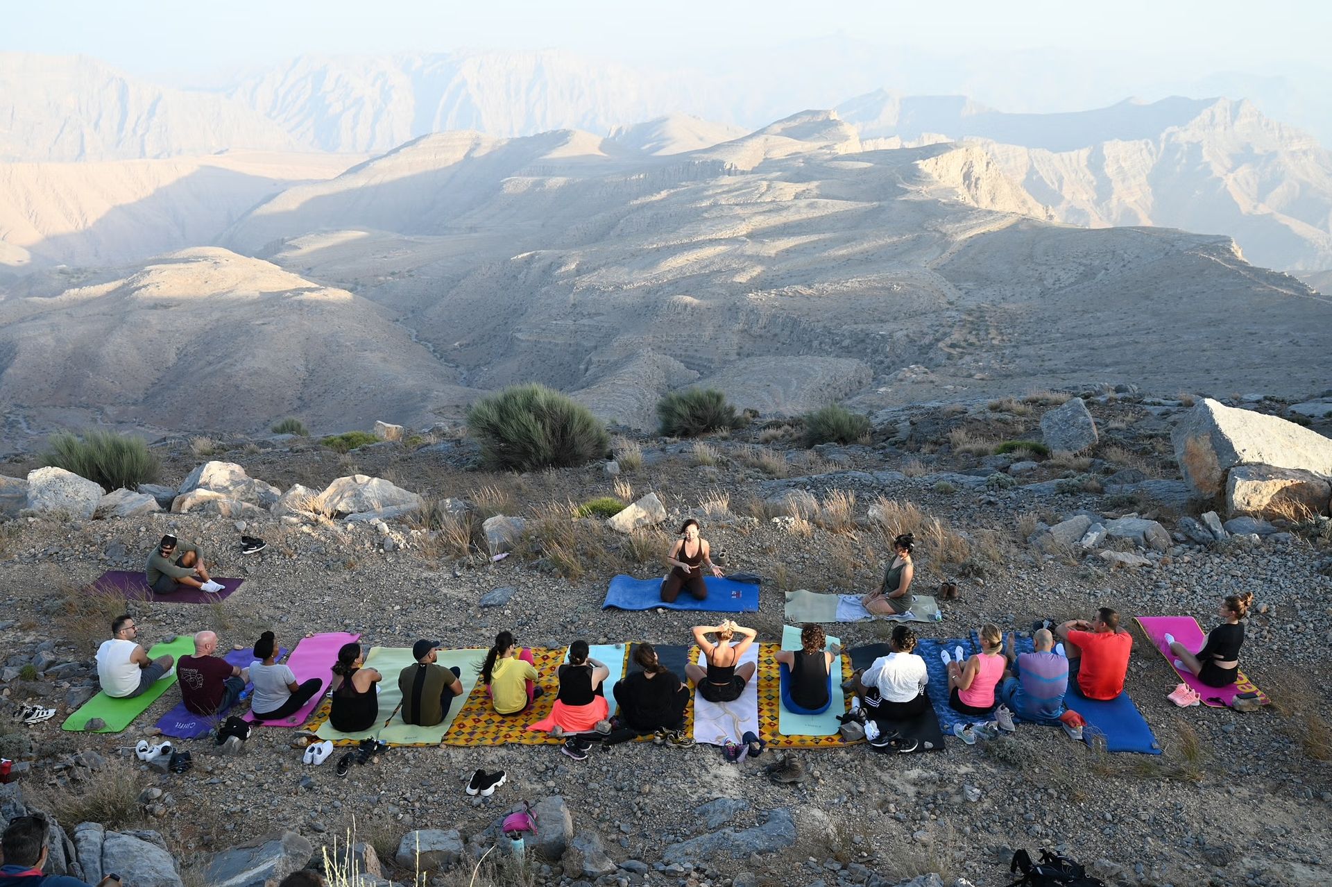 Group doing yoga on a mountain ledge at sunrise, overlooking a vast rocky valley and distant peaks in Jebel Jais.