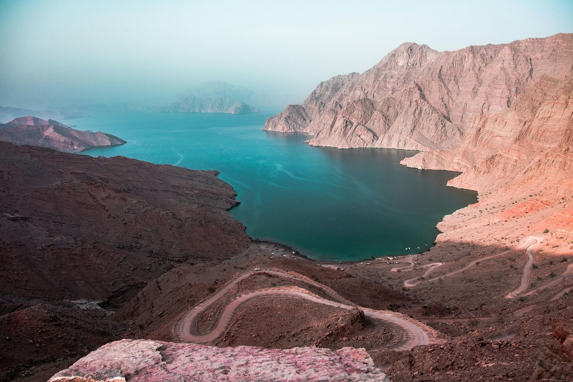 Rocky canyon overlooking a turquoise bay with winding dirt road and hazy mountains in the distance in Khasab Musandam.