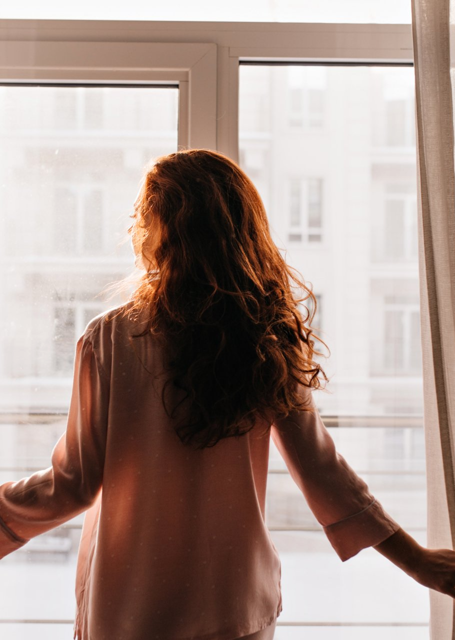 a woman touching sheer curtains looking outside the window