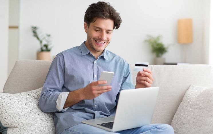 Um homem está sentado em um sofá usando um laptop e um telefone celular.