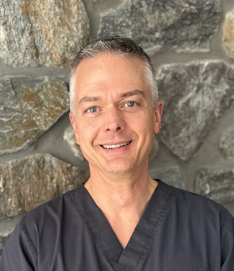 A man in a scrub top is smiling in front of a stone wall.