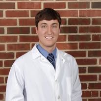 A young man in a lab coat and tie is standing in front of a brick wall.