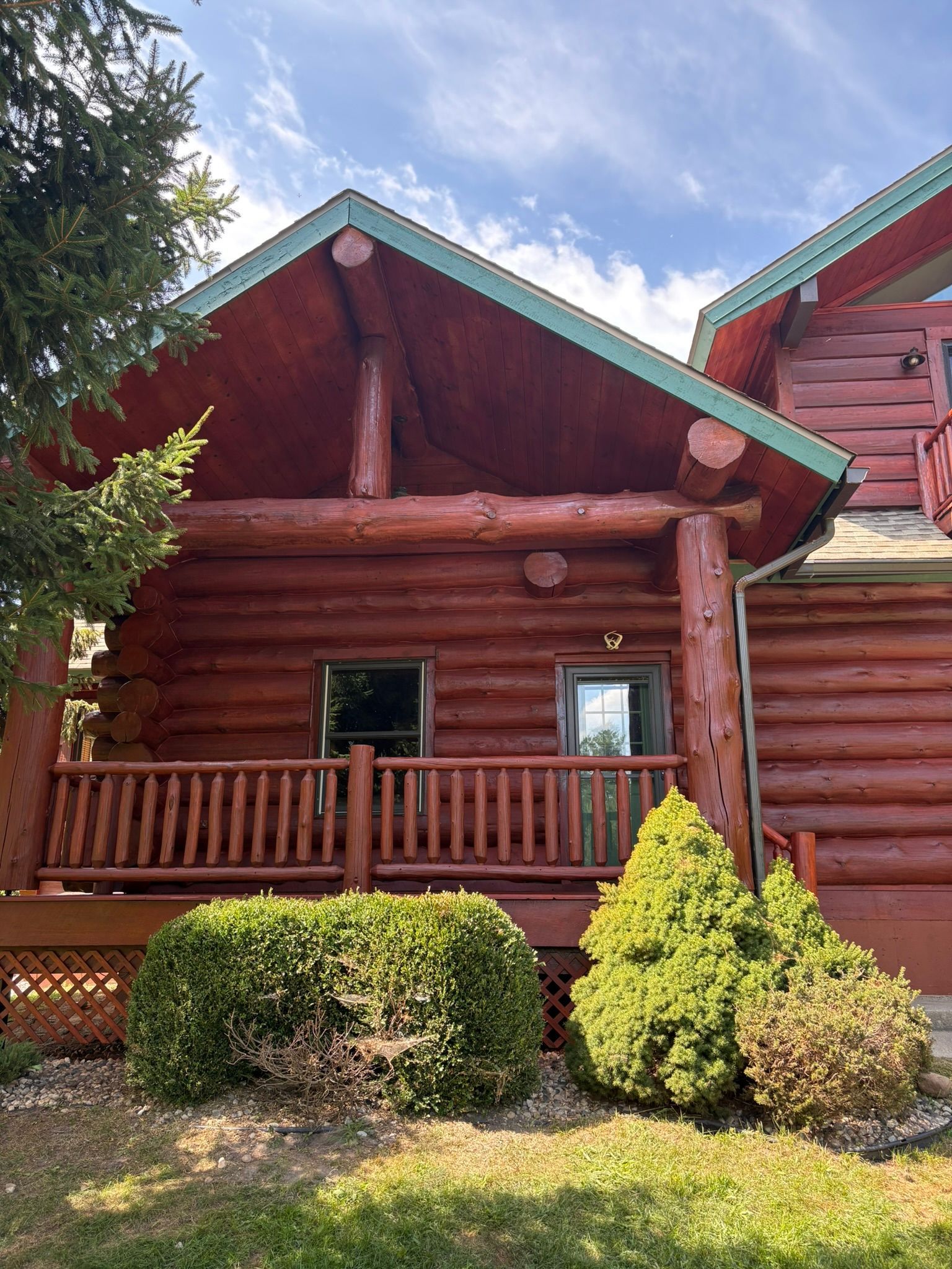 Log cabin with red log siding, green roof trim, and a small porch surrounded by shrubs.