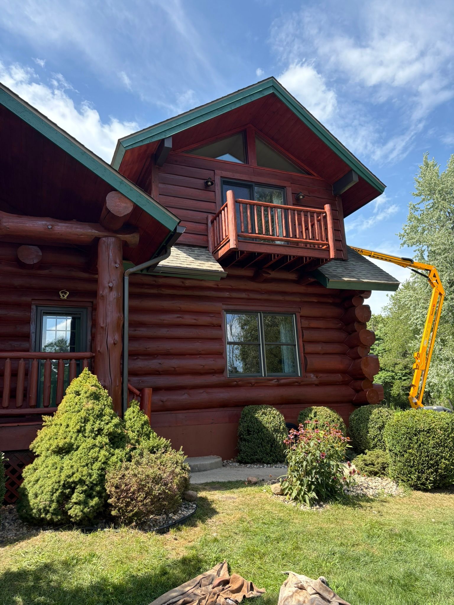 Log cabin with green trim and balcony, surrounded by shrubs under a bright blue sky.