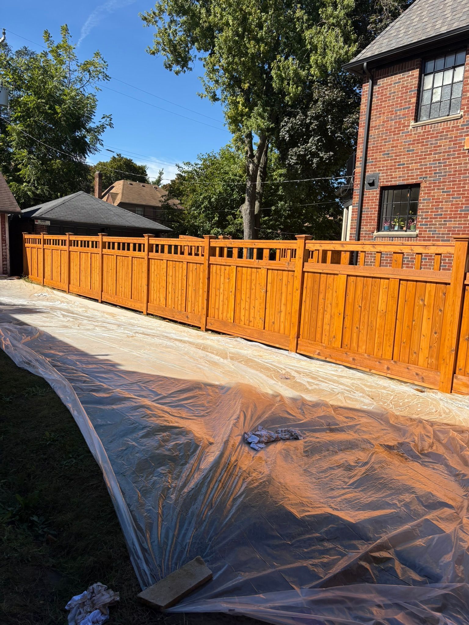 Orange wooden fence along a paved driveway beside a brick house, with trees and blue sky in the background