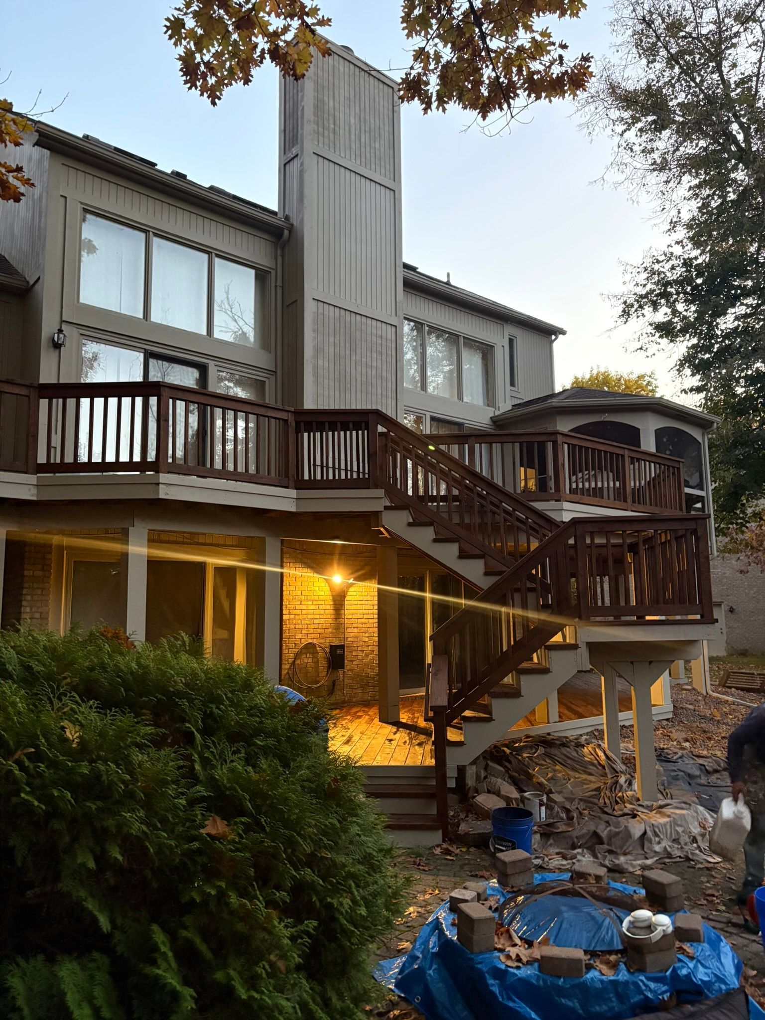 Two-story house with a large wooden deck, staircase, and lit patio beside a stone-lined pool at dusk