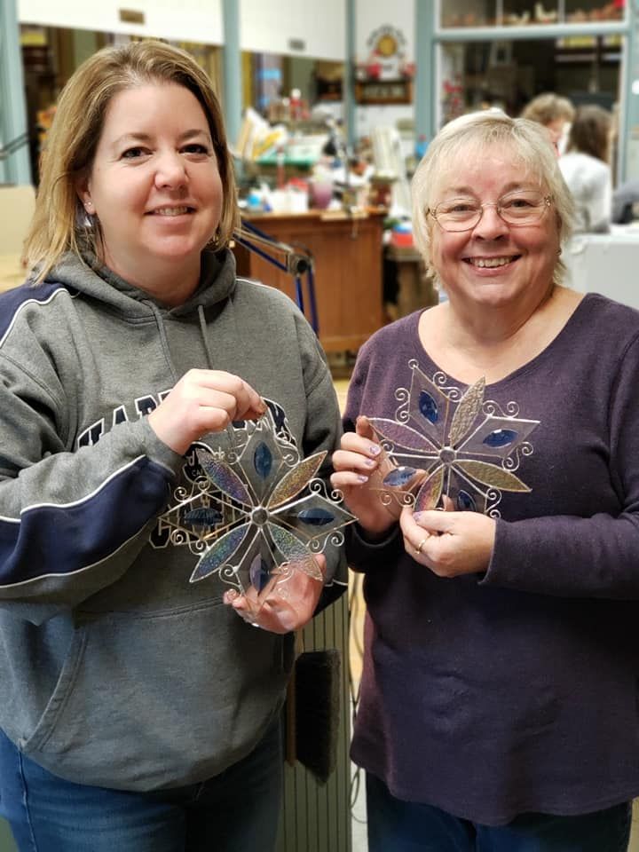 Two ladies  showing off their stained glass workshop projects-clear beveled snowflakes with shades of blue. 