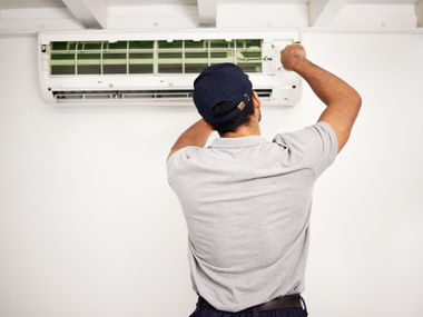 A gray residential air conditioning condenser unit sits on a concrete patio against a gray wall.