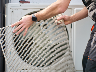 Three air conditioning units and a smaller heat pump sit on a concrete pad against a brick wall next to a metal door.