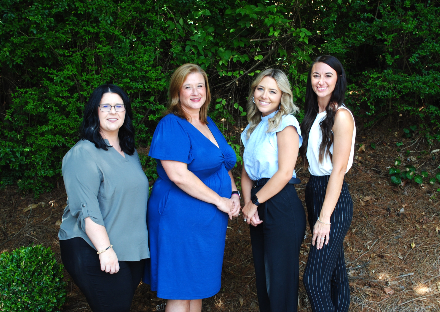Four women standing outdoors in front of green bushes, posing and smiling at the camera.