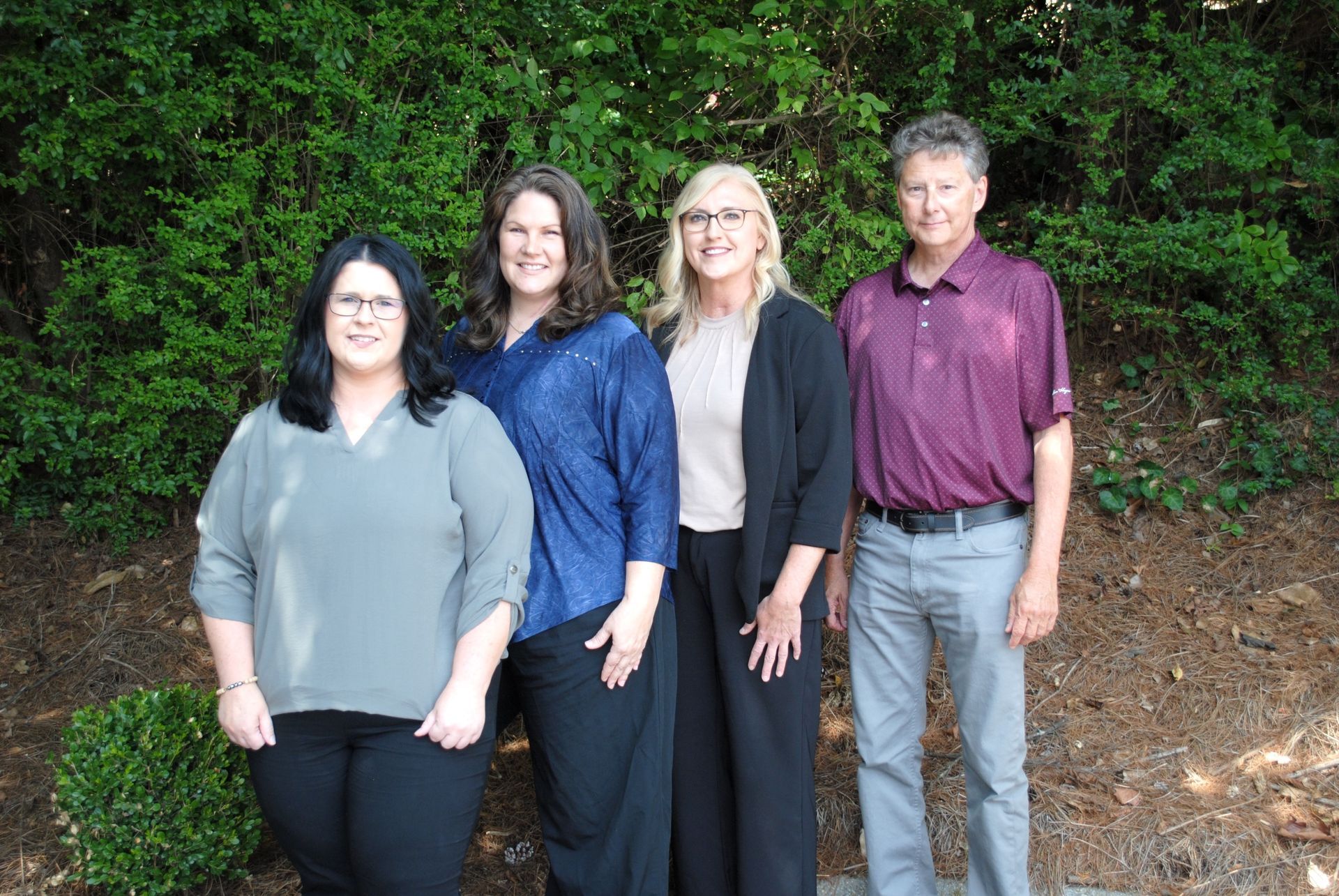 Four people standing outdoors in front of green bushes, smiling at the camera.