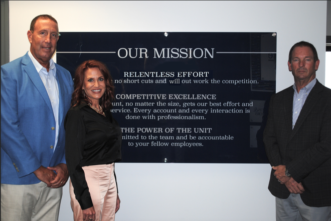 Three people stand beside an “OUR MISSION” wall sign in an office lobby.